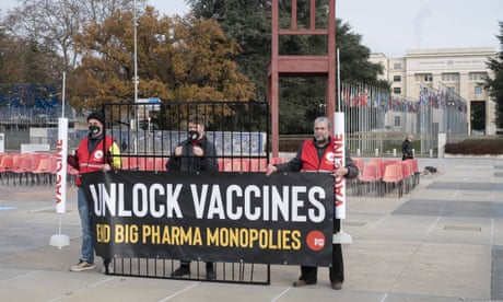 Three men stand behind a makeshift jail grate. The two on either end are holding enlarged needles with the word 'vaccine' on them. A sign in front of them says 'Unlock vaccines. End big pharma monopolies.'