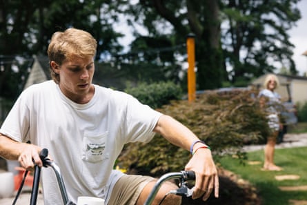 A man wearing a white T-shirt adjusts his bike outside, his mother looks on in the background