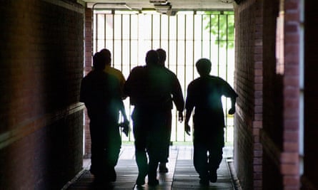 A group of people with their backs to the camera walking down a corridor, silhouetted by the light coming through a barred gate at the end