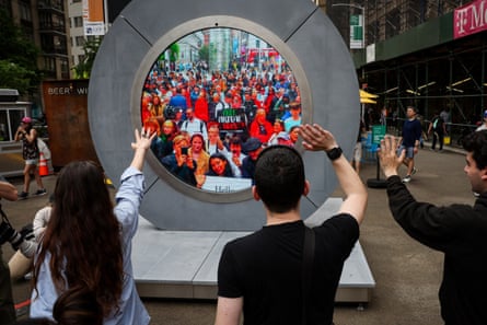 Three people wave at a circular grey structure with a screen of a crowd of people
