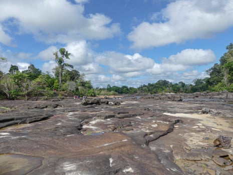 The stony bed of a wide river bordered by jungle, with people seen 100 metres away where there remains water