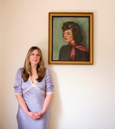 Daisy Buchanan wearing a lilac, lace-trimmed dress standing with her hands clasped in front of her next to a painting of a dark-haired woman wearing a red neckerchief