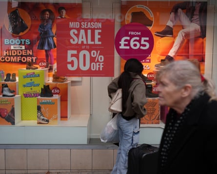 People walk past a shop window with large red “Mid Season Sale” signage advertising discounted boots and shoes