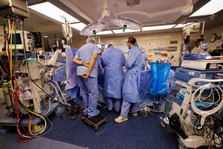 Surgeons in blue surgical gowns, with back to the camera, are gathered around the patient during the procedure