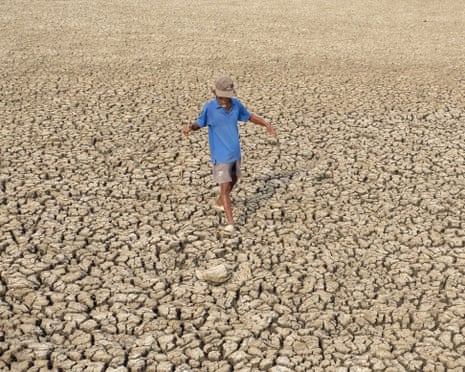 A person walks barefoot across a dry, deeply cracked earth surface extending to the horizon