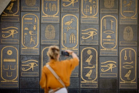 A woman takes a picture of an entrance covered in large golden hieroglyphs