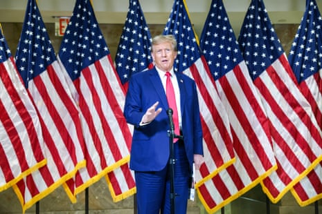 A man in a blue suit and red tie speaks in front of a row of American flags