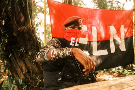Commander Yerson with a red and black bandana covering his nose and mouth standing in front of large ELN flag
