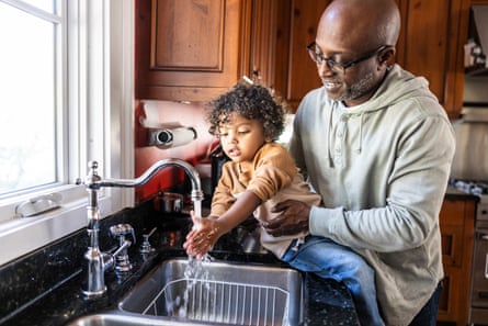 A father helping a toddler wash hands in a kitchen sink.