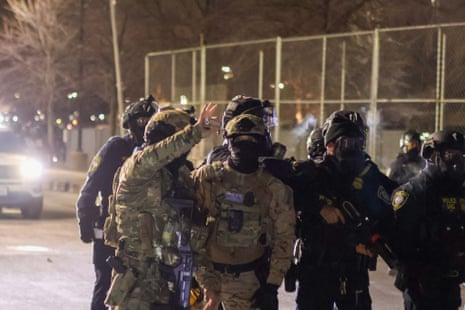 A federal agent in camoflauge uniform and helmet holds up a flashbang grenade while standing with other law enforcement officers in black riot gear and one other federal agent in camoflauge gear. They are standing on a road under a dark sky with a car shining a headlight behind them.