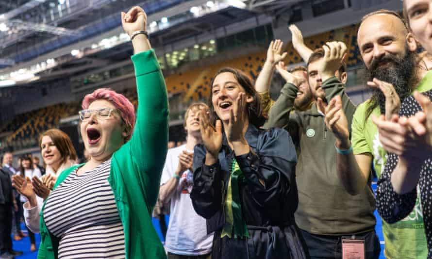 Large group of people clap and cheer. Woman wears bright green cardigan; others also wearing green