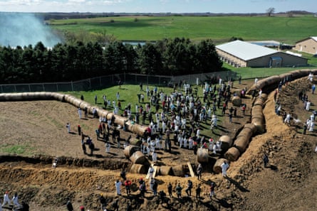 Aerial view of dozens of people crossing a trench and pushing aside round hay bales, with fence in background on other side of one-story barn.