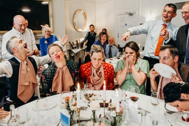 A group of friends at a wedding sit around a table, some with dessert around their mouths, while others look on and point and laugh