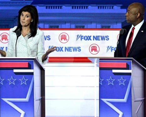 A white woman and a Black man stand at neighboring lecterns on a brightly lit stage. The woman is speaking, with both palms raised, as the man listens.