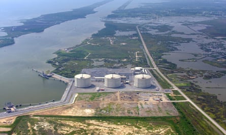 Aerial view of three white silos in a row on an asphalt aprob, surrounded by flat green land and a water channel, beyond which is a great expanse of water.
