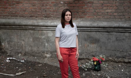 Liliia Yakovleva, wearing a T-shirt and red trousers, standing against a brick wall, with dirt and some rubble on the floor, and a vase of flowers