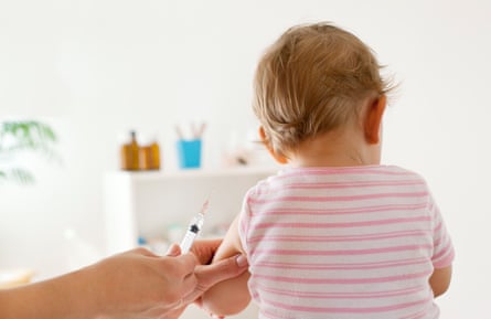 Back view of a baby wearing a pink striped shirt receiving a vaccination.
