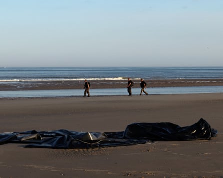 Three police officers walk across the sands in the background behind a punctured black inflatable boat