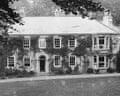 Black and white picture of large two-storey country house with pram bottom right and woman looking out of window
