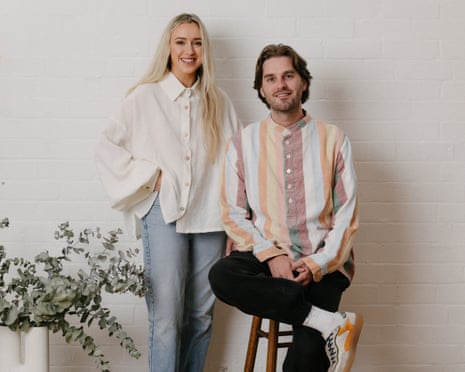 Image 4: Joel sits on a stool with one leg crossed over his knee and wearing a striped linen shirt. Amy, with long blonde hair worn loose, stands next to him wearing a white linen shirt.