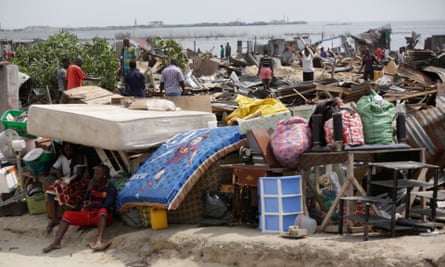 Africans sit and stand around in the ruins of their demolished homes with their belongings in piles around them and the sea in the distance.