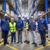 Six people including Denis Mukwege and Ellen Sirleaf in hard hats stand in a warehouse with blue racking