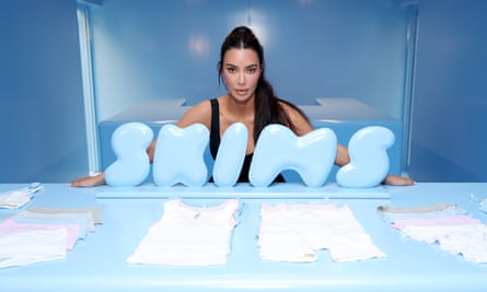 Kim Kardashian poses for a photo behind an upright 3D rendering of the pale blue Skims logo, with a table displaying various Skims garments in front of it
