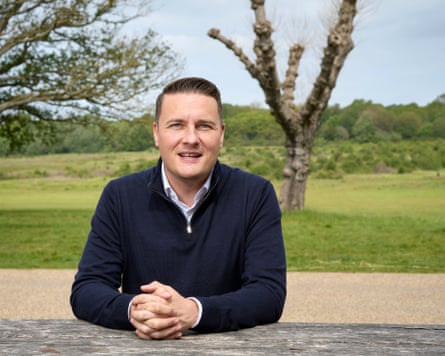 Wes Streeting leans on a wooden fence at a country park in Essex, with trees and green space behind him.