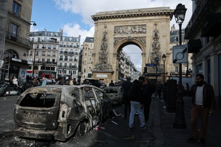 People stand by a burnt-out car in a Paris street with debris in the road and a stone arch in the background.