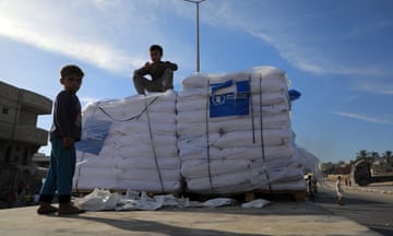 A child sits on a pile of aid parcels while another standing in front of it looks at the camera