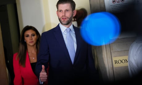 A white man with a scruffy beard, dark suit, light blue shirt and blue tie gives a thumbs up outside a courtroom door, with a woman with long brown hair and red blazer behind him.