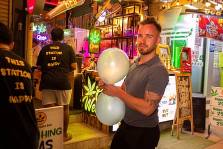 A man holds two balloons outside a weed shop where two shop workers wear shirts saying: ‘This station is happy’