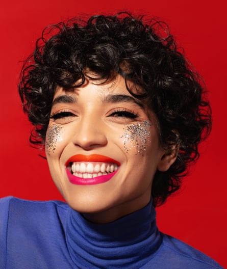 A smiling woman’s face with glitter on her cheeks, orange lipstick on her upper lip and pink lipstick on her lower lip