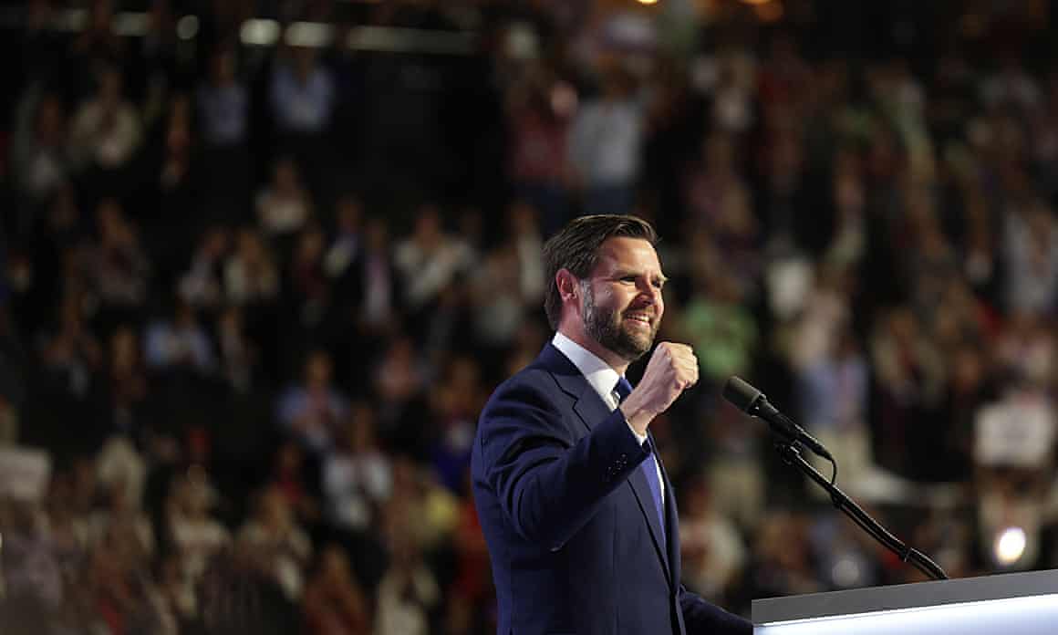 A smiling JD Vance stands before a microphone with his hand raised with a large crowd blurred in the background at the Republican National Convention in Milwaukee, Wisconsin.