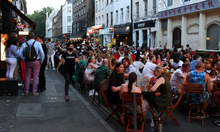 A waitress in a T-shirt, jeans and mask walks down Frith Street, where many people are eating at tables in the street