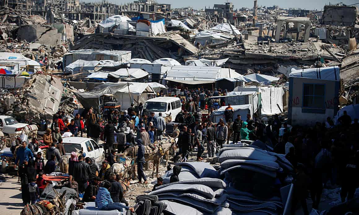 Crowds of people amid Tarpaulin-covered structures in front of bombed-out remain of northern Gaza