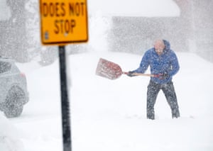 A man in blue padded coat uses a show shovel as snow falls. A road sign saying 'Does not stop' is in the foreground