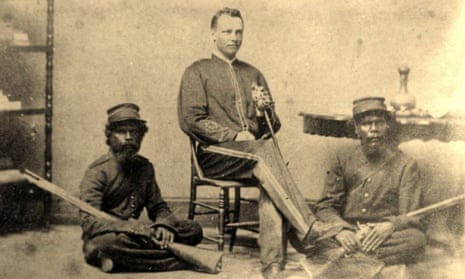 Reginald Uhr as a young man wearing police uniform, seated on a wooden chair. Seated cross legged on the ground are two Aboriginal troopers.