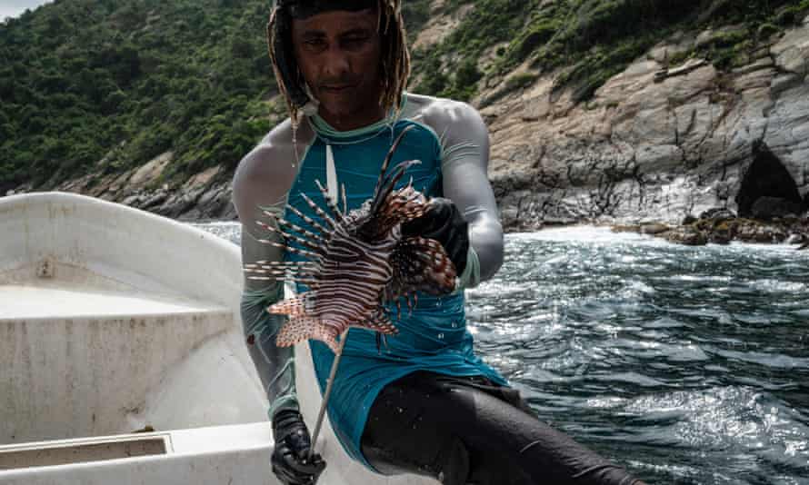 A spearfisher sits on the side of a boat holding out a lionfish’s poisonous spiky fins 