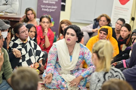 A dozen or so people gather round a person sitting cross-legged on the floor