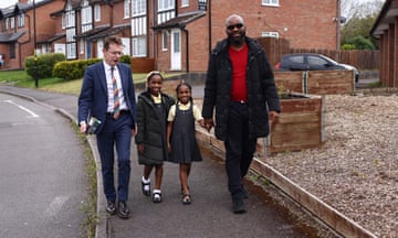 Andy Street in a suit and tie, holding a clipboard, walks down a residential street with two smiling Black schoolgirls, one holding hand with her father, who is wearing sunglasses, smiling and holding a clipboard