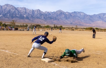 A crouching man in a baseball uniform holds up a glove as another man dives in the dirt to the base at his feet, on a dusty field with mountains beyond.
