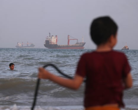 Children play in the water, with vessels sit offshore in the distance