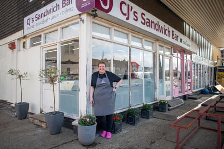 A woman in an apron stands outside a cafe building.
