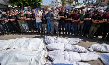 A line of people stand with their arms crossed with bodies wrapped in white material laid before them.