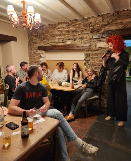 A man wearing a red wig and a black dress standing with a microphone in front of customers sitting at tables