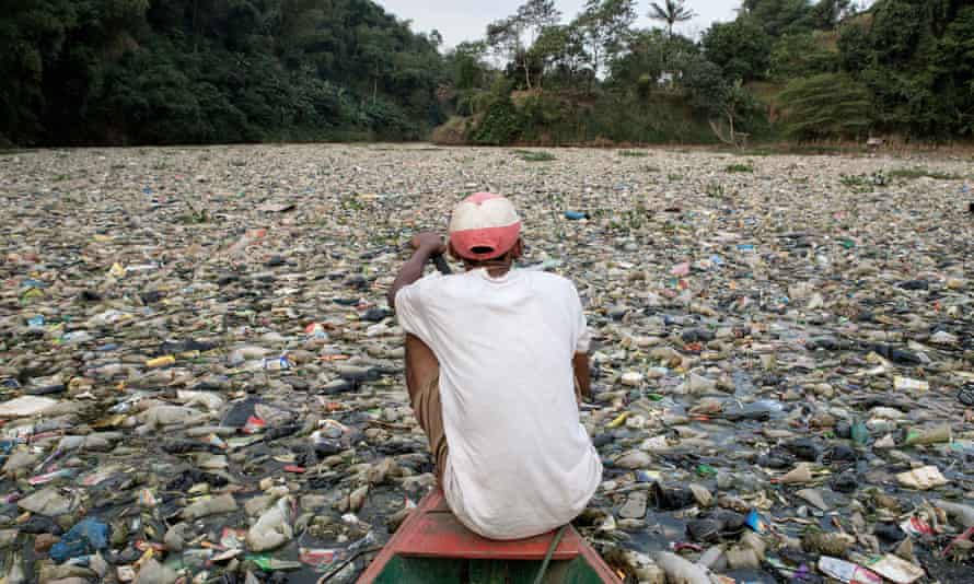 A man in the bow of a small boat paddles through a floating mat of rubbish that covers the surface of a wide river