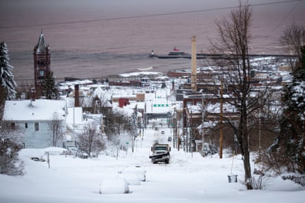 A snow plow travels up a street in a residential neighborhood with a large lake in the background.