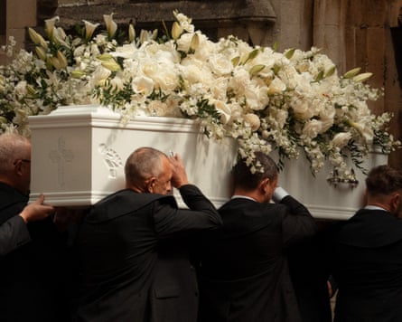 Men in black carry the white coffin, which is covered in white flowers