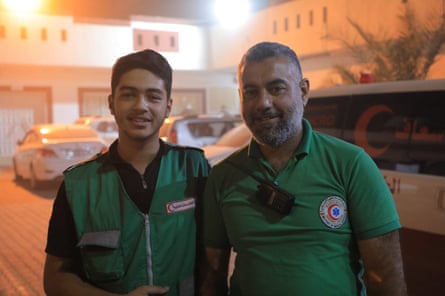 A smiling young man in a green paramedic vest stands next to an older man wearing a green ambulance service t-shirt.